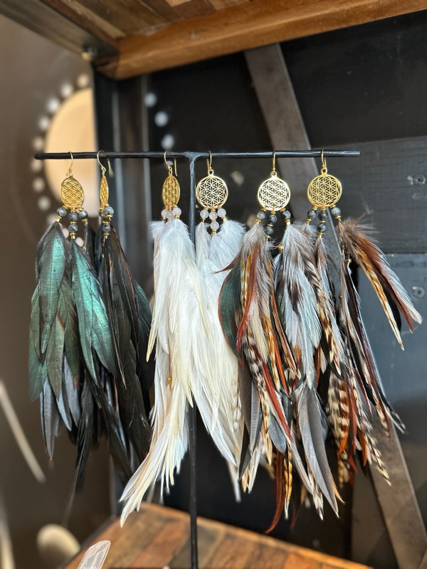 Feather earrings in white, teal and brown hanging from a rail, each with a gold Flower of Life charm