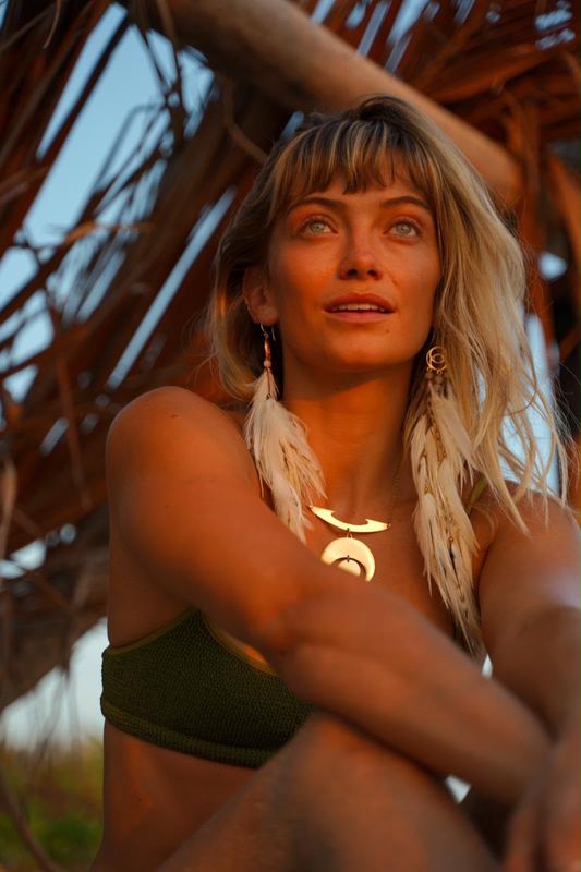 A woman in warm golden light under a thatched palm roof, wearing long white feather earrings with gold charms