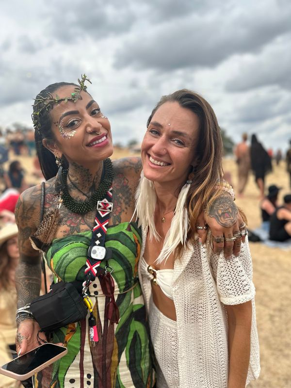 Two friends arm in arm at a festival under a cloudy sky, one wearing long white feather earrings