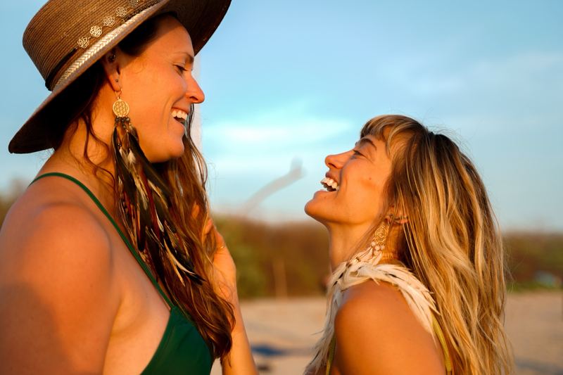 Two women laughing on a beach in golden evening light, one wearing a long feather earring with a gold charm