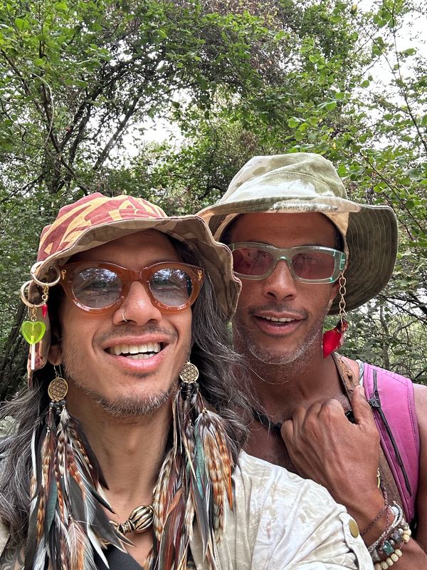 Two friends in bucket hats and tinted sunglasses among trees, one wearing long brown, green and light feather earrings with gold charms