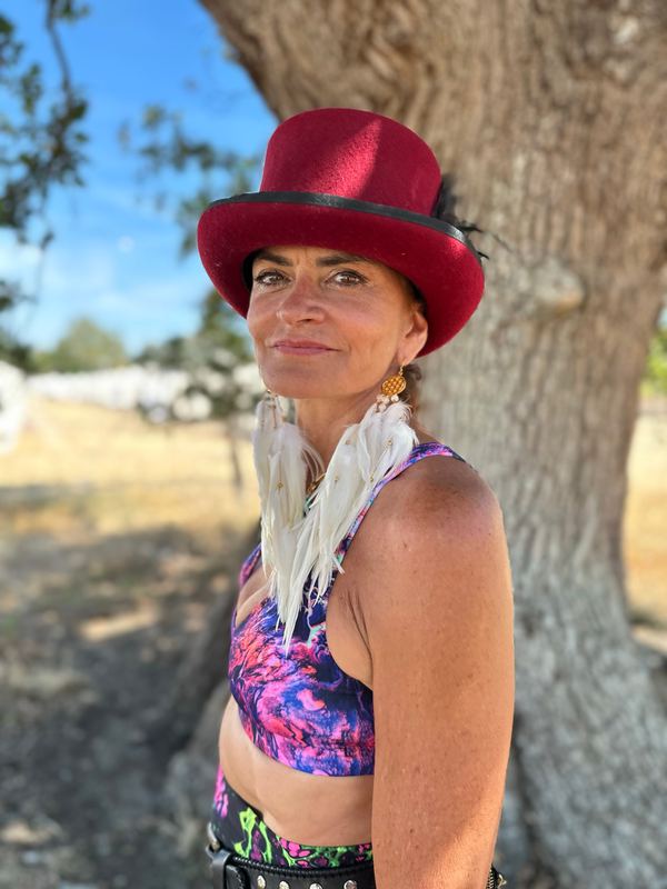 A woman in a red top hat leaning against a tree, long white feather earrings resting on her shoulders