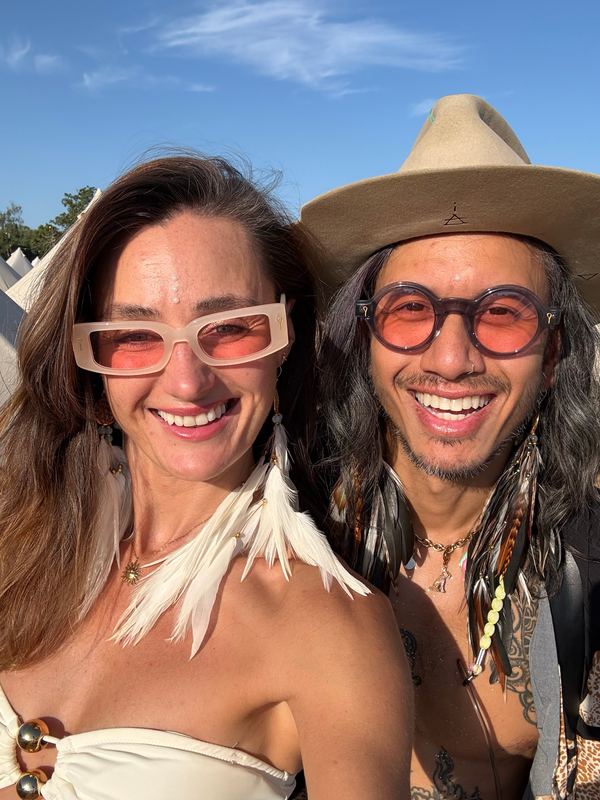 Two friends smiling under a blue sky, one wearing white feather earrings