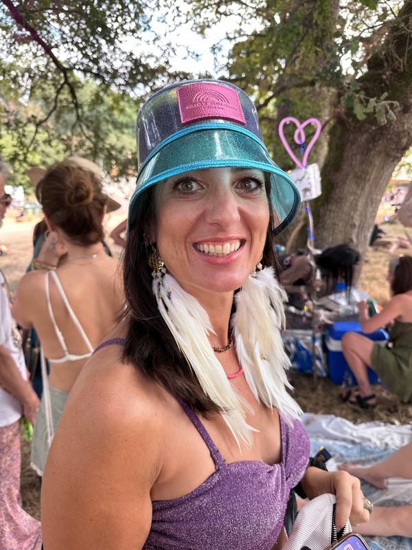 A woman in a glittering turquoise bucket hat at a festival, wearing long white feather earrings