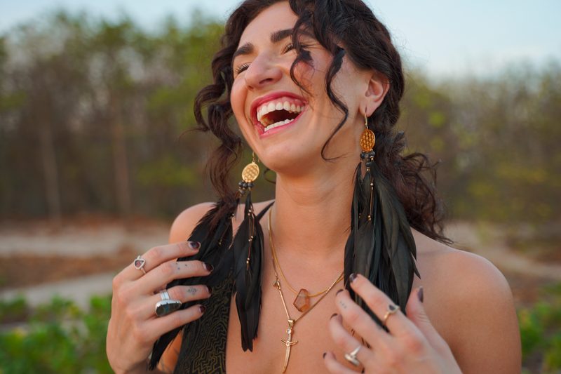Laughing with head tilted back on a beach path, black feather earrings with gold Flower of Life charms in her hands