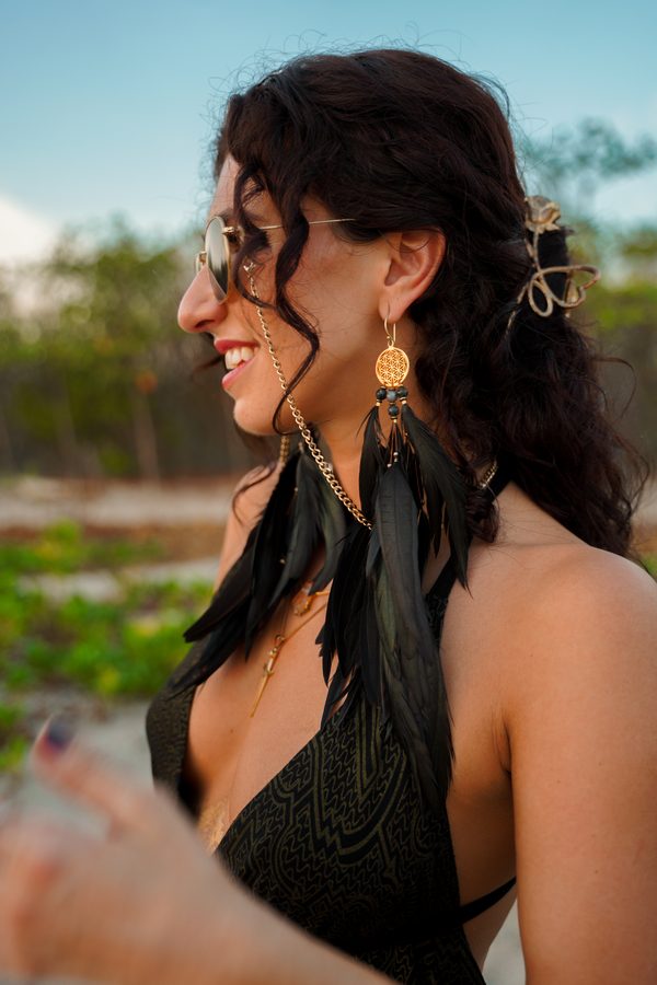 Smiling in profile in round sunglasses, a long black feather earring with a gold Flower of Life charm